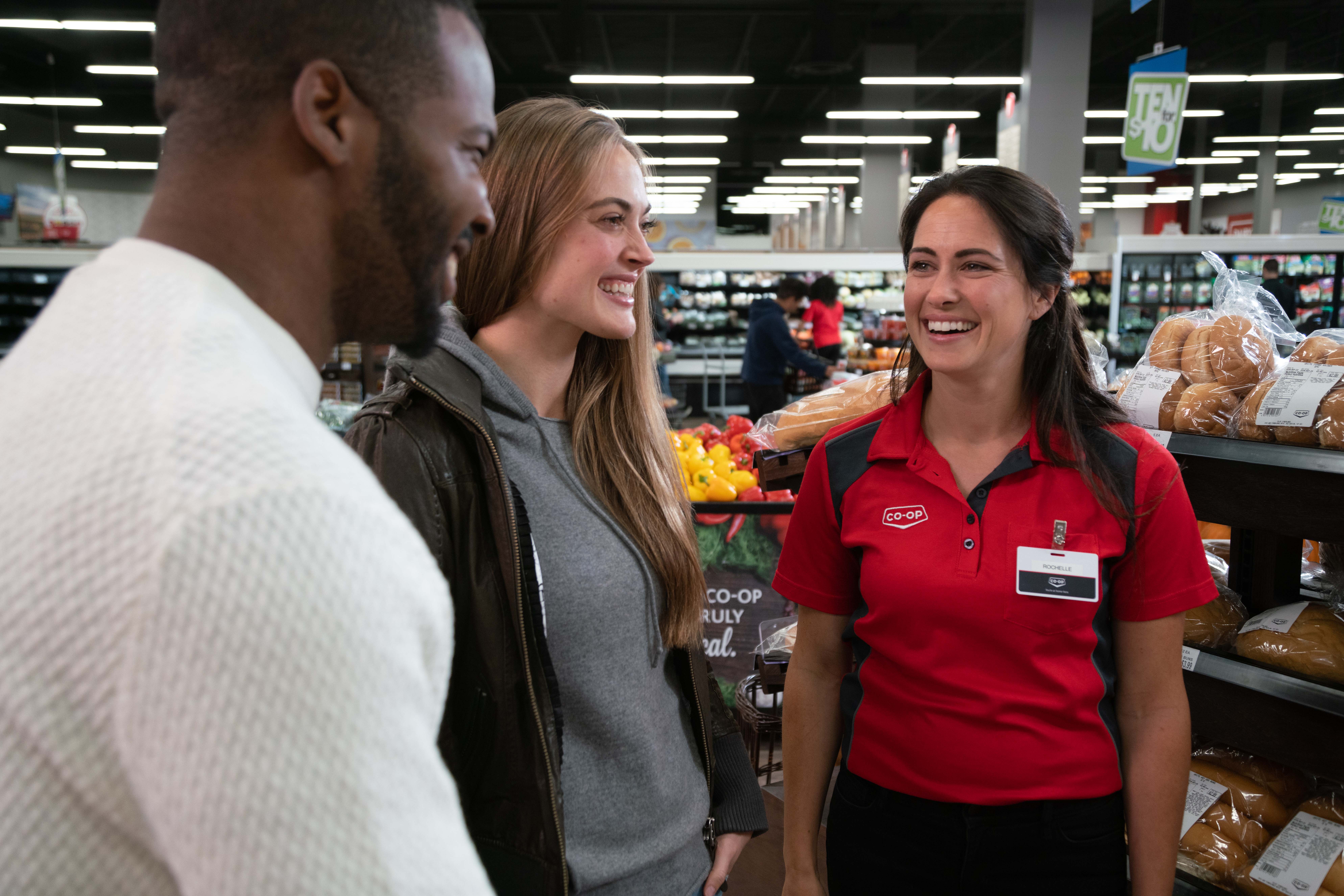 Three smiling individuals standing in a semi-circle in a grocery store bakery aisle.